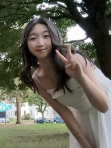 A person in a white dress stands outdoors, smiling and holding up a peace sign with one hand. Trees, grass, and houses are visible in the background.