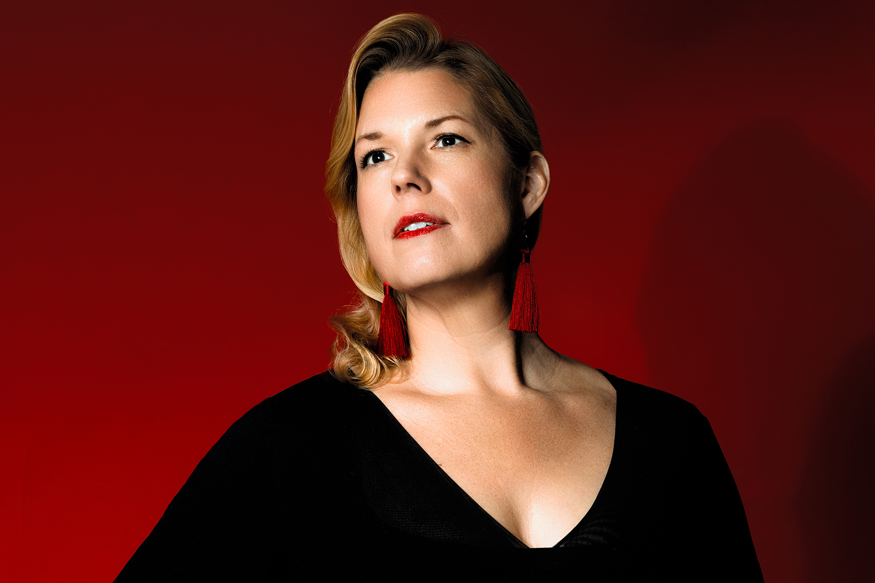 A woman with blonde hair, red tassel earrings, and a black top poses against a solid red background, looking slightly upward.
