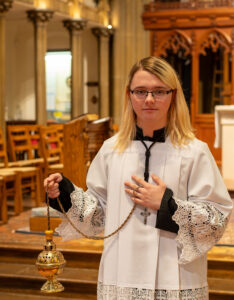 A person in a white robe holds a censer inside a church, standing near wooden pews and an altar.