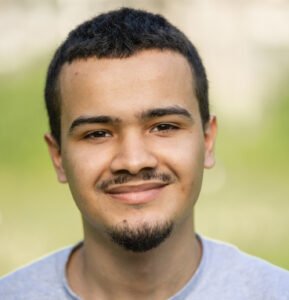 Person with short dark hair and a light mustache and goatee, wearing a gray shirt, smiling outdoors with a blurred green background.
