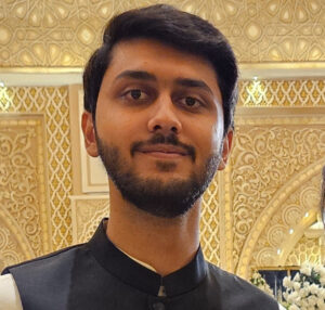 A person with short dark hair and a beard stands indoors in front of an ornate, gold-patterned background, wearing a black and white outfit.