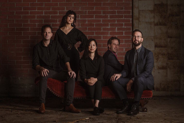 Five people sit and pose on a red velvet couch against a brick wall, dressed in dark formal clothing, looking at the camera.