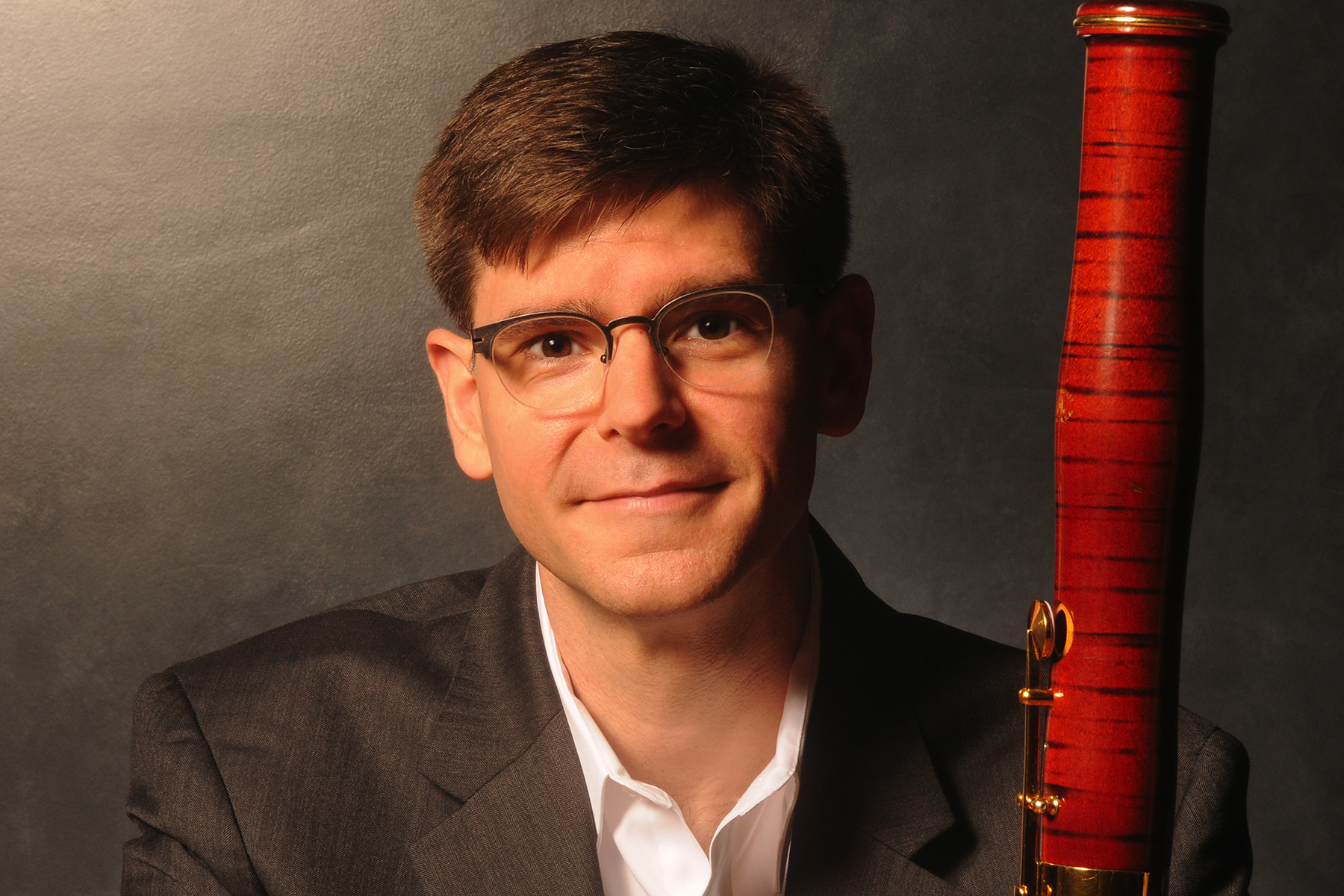 A man in a suit jacket and glasses holding a bassoon.