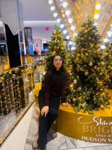 A person sits beside decorated trees with golden lights and ornaments inside a brightly lit shopping center.