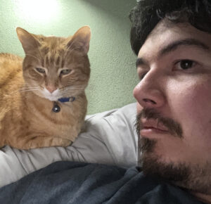 A person lies on a bed next to an orange cat wearing a collar, both looking toward the camera.