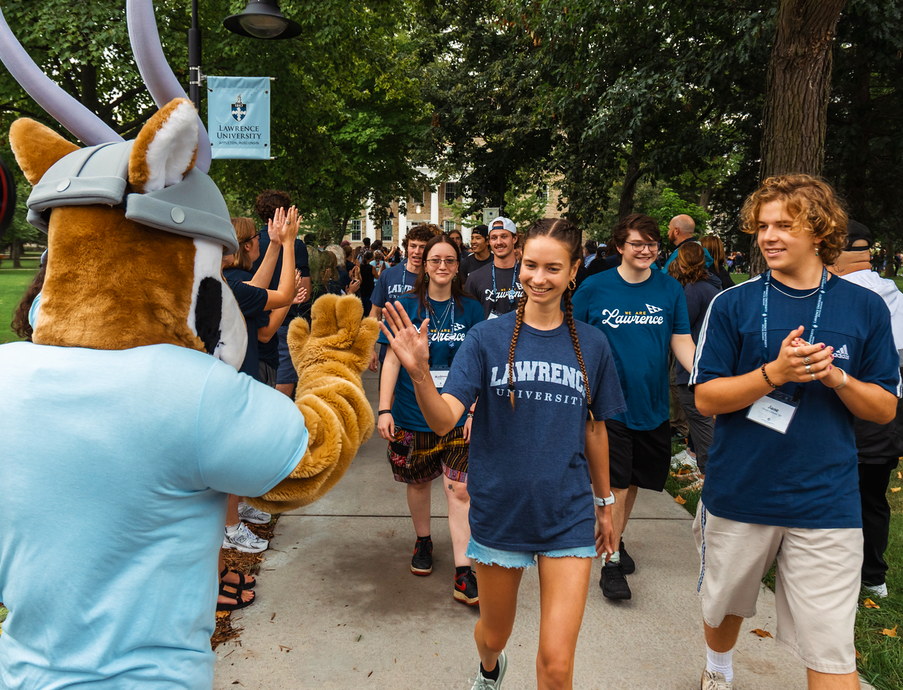 A group of students in Lawrence University shirts walk on a sidewalk, high-fiving a Viking mascot. Trees and campus buildings are visible in the background.