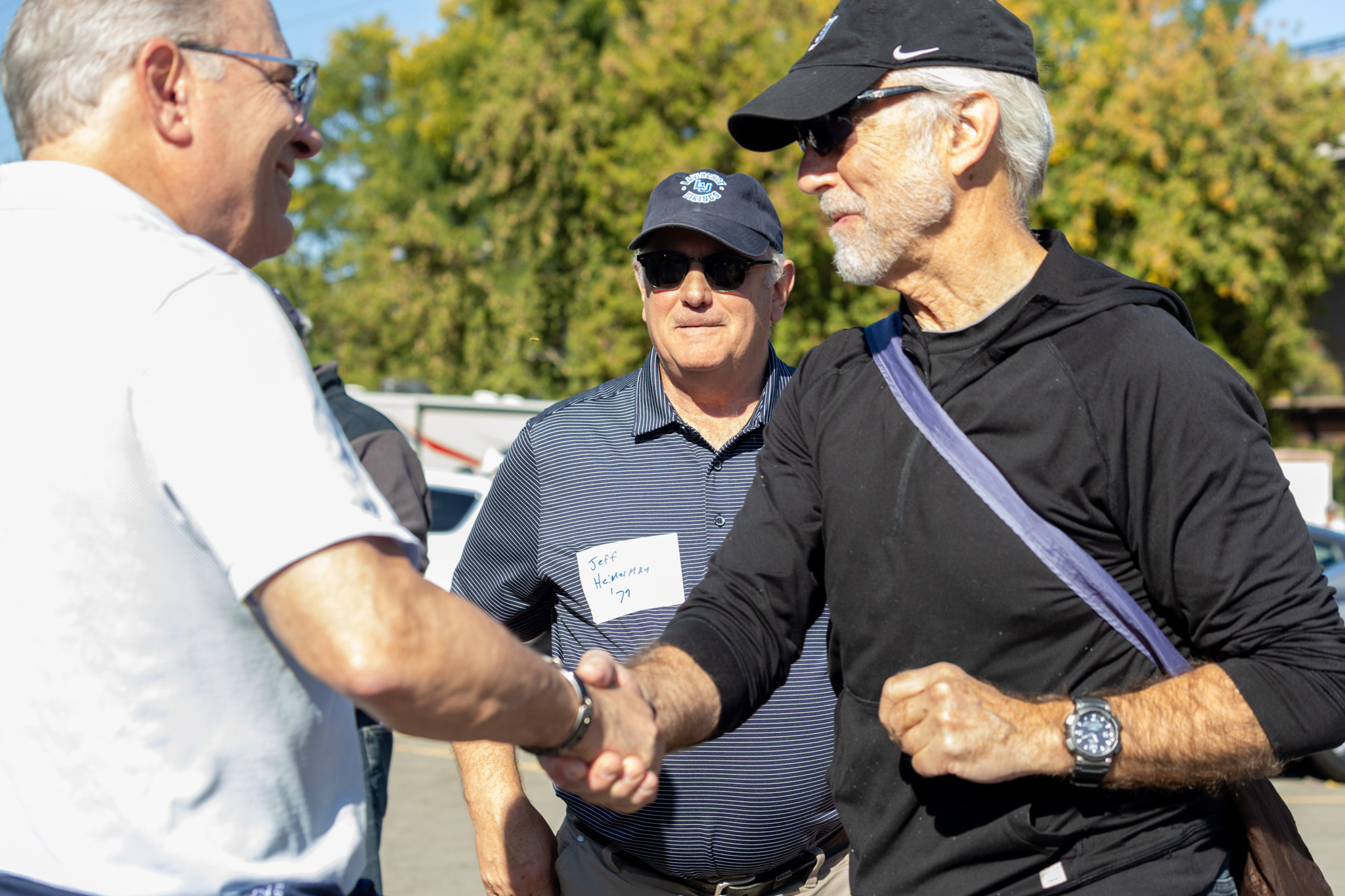 Two men shaking hands outdoors while a third man in a blue polo shirt and name tag watches.
