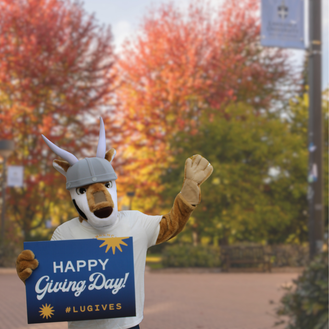 A university mascot wearing a Viking helmet holds a sign that reads HAPPY Giving Day! #LUGIVES and waves.