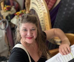A person smiling in front of a harp while holding a sheet of music.